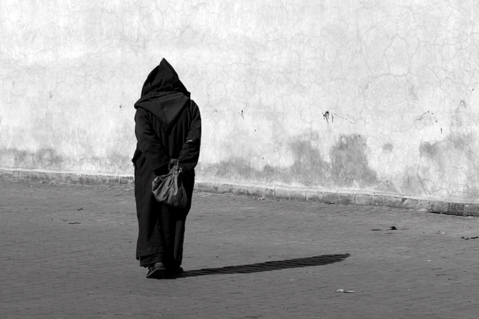 Back view of a dark hooded figure with hands clasped behind back, casting a long shadow