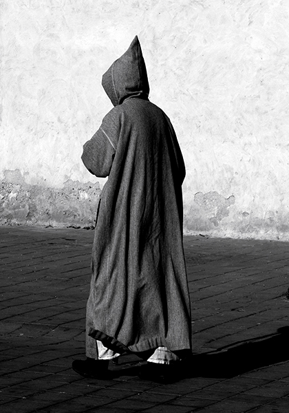 Back view of a dark hooded figure walking in front of a bare stone wall
