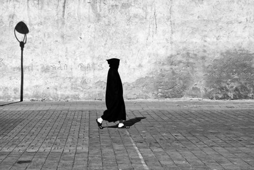 Person dressed all in black except for white socks walking past a bare stone wall with the shadow of a lamp post